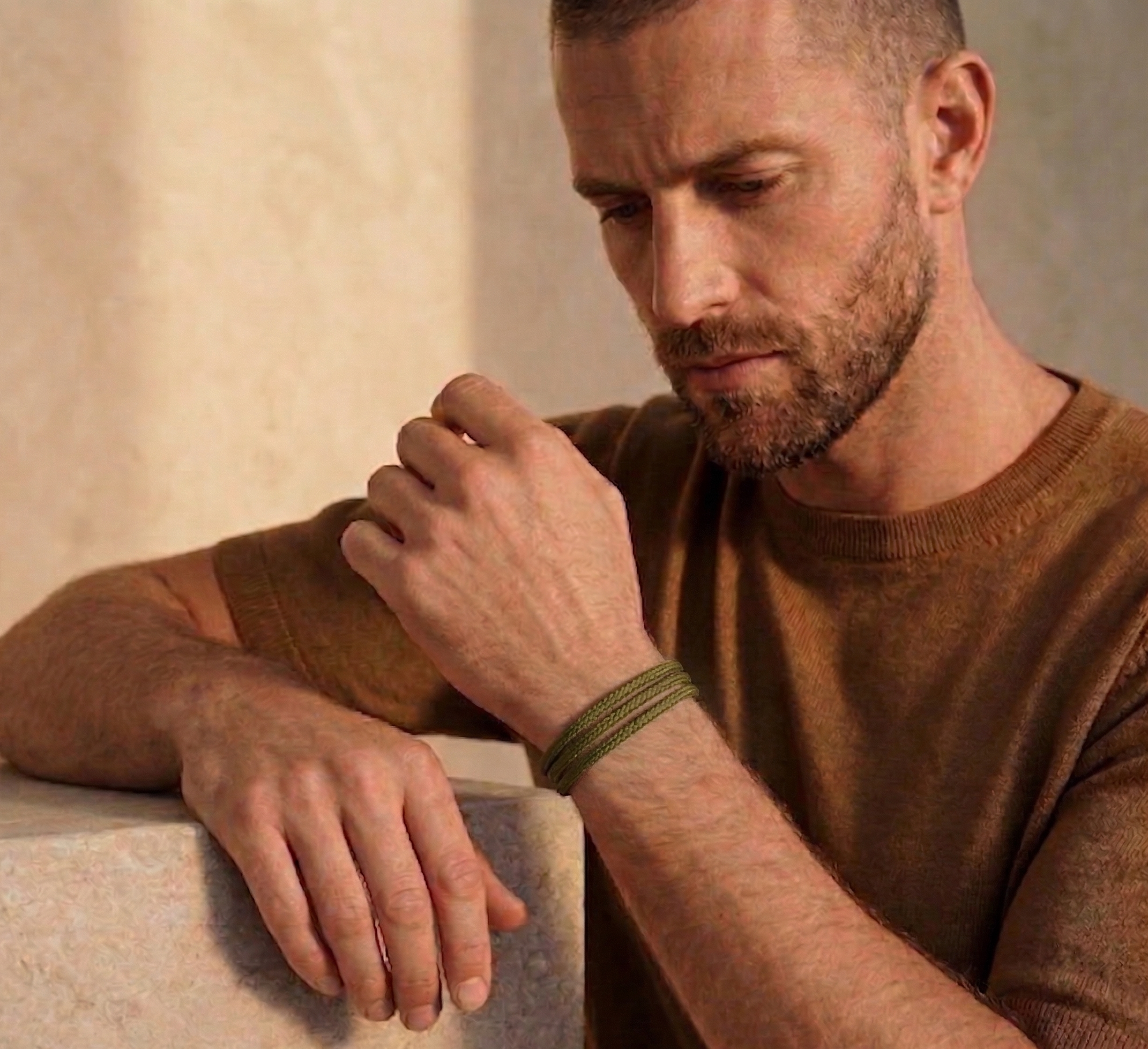 Man wearing a brown sweater and green bracelet, leaning on a stone surface.